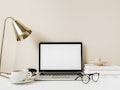 Blank screen laptop. Home office desk table workspace with coffee, lamp, glasses, notebook on beige ...