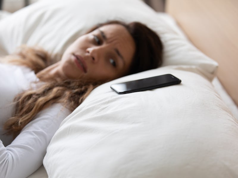 Close up focus on mobile phone is on pillow, frightened woman lying in bed looking on smartphone fee...
