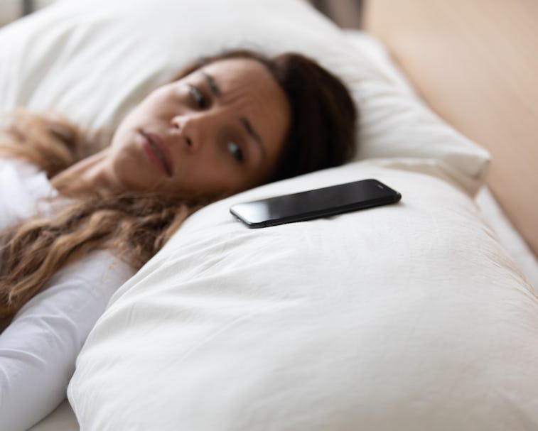 Close up focus on mobile phone is on pillow, frightened woman lying in bed looking on smartphone fee...