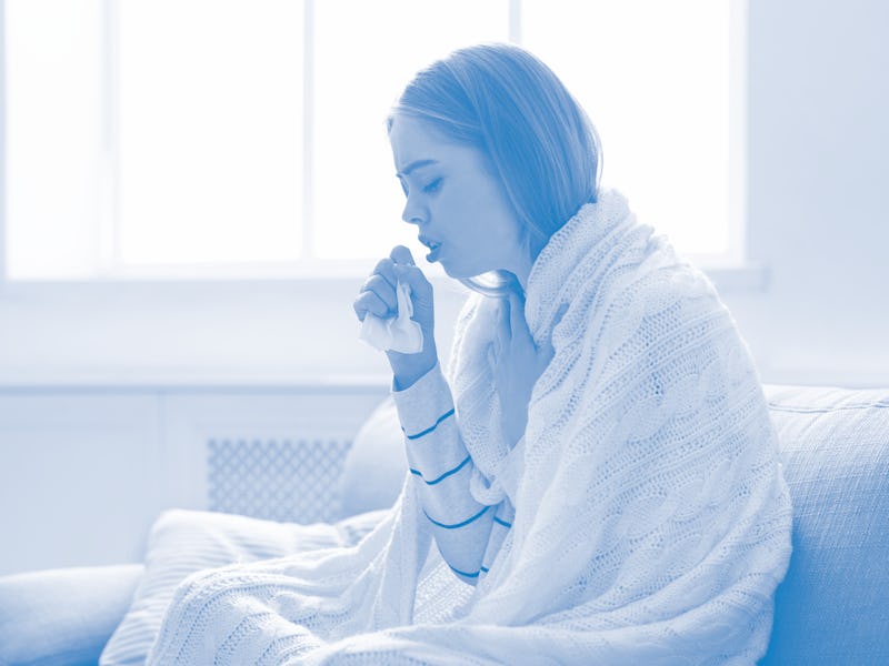 A woman is seen wrapped in a blanket, coughing into her hand. She is holding a tissue and looks ill.