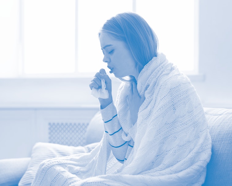 A woman is seen wrapped in a blanket, coughing into her hand. She is holding a tissue and looks ill.