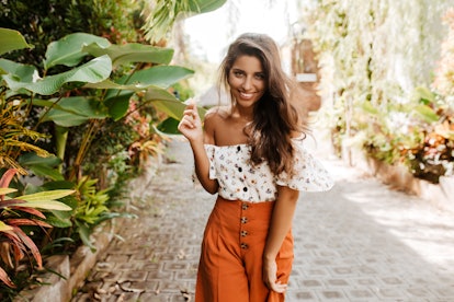Positive woman in orange wide shorts and white top smiles coquettishly, holding small flower in her ...