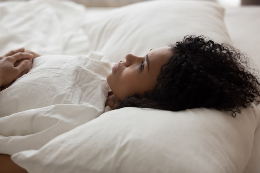 Exhausted African American woman with open eyes lying in bed under white blanket, tired girl with cu...