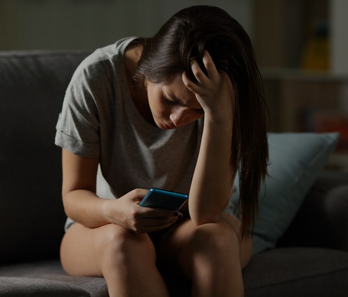 Sad teen being victim of cyber bullying online sitting on a couch in the living room at home