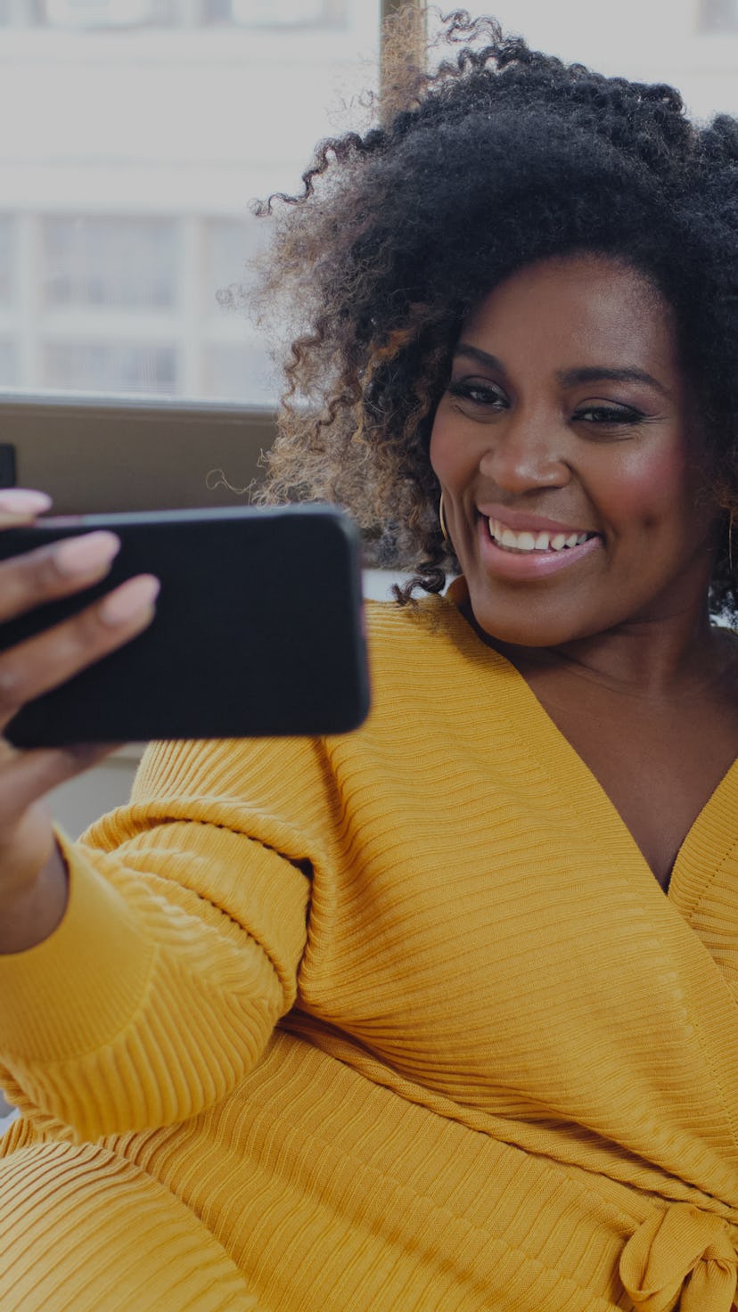 A happy woman is seen smiling at a smartphone while holding a glass of champagne. She is wearing a b...