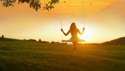 GOLDEN LIGHT SILHOUETTE: Unrecognizable girl in white dress swaying on a tree swing on peaceful even...