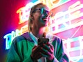 Image of beautiful young caucasian woman in 3D glasses holding smartphone over multicolored neon tex...