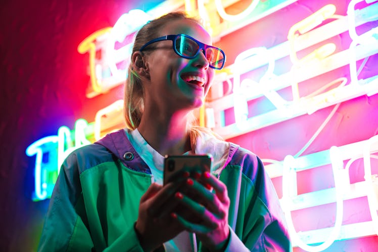 Image of beautiful young caucasian woman in 3D glasses holding smartphone over multicolored neon tex...