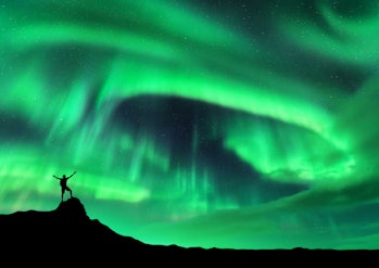 Aurora borealis and silhouette of a man with raised up arms on the mountain peak. Lofoten islands, N...
