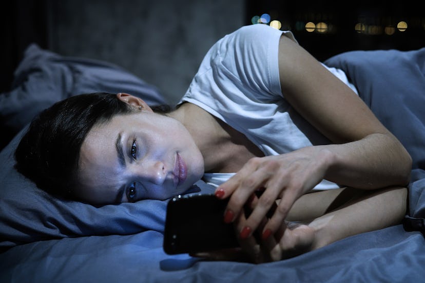 Smartphone addiction. Young tired female looking at her mobile phone screen, lying in bed late at ni...