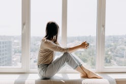 Young woman looking through the window with a city view, sitting on a windowsill, drinking coffee or...