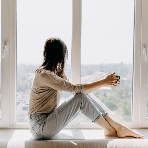 Young woman looking through the window with a city view, sitting on a windowsill, drinking coffee or...