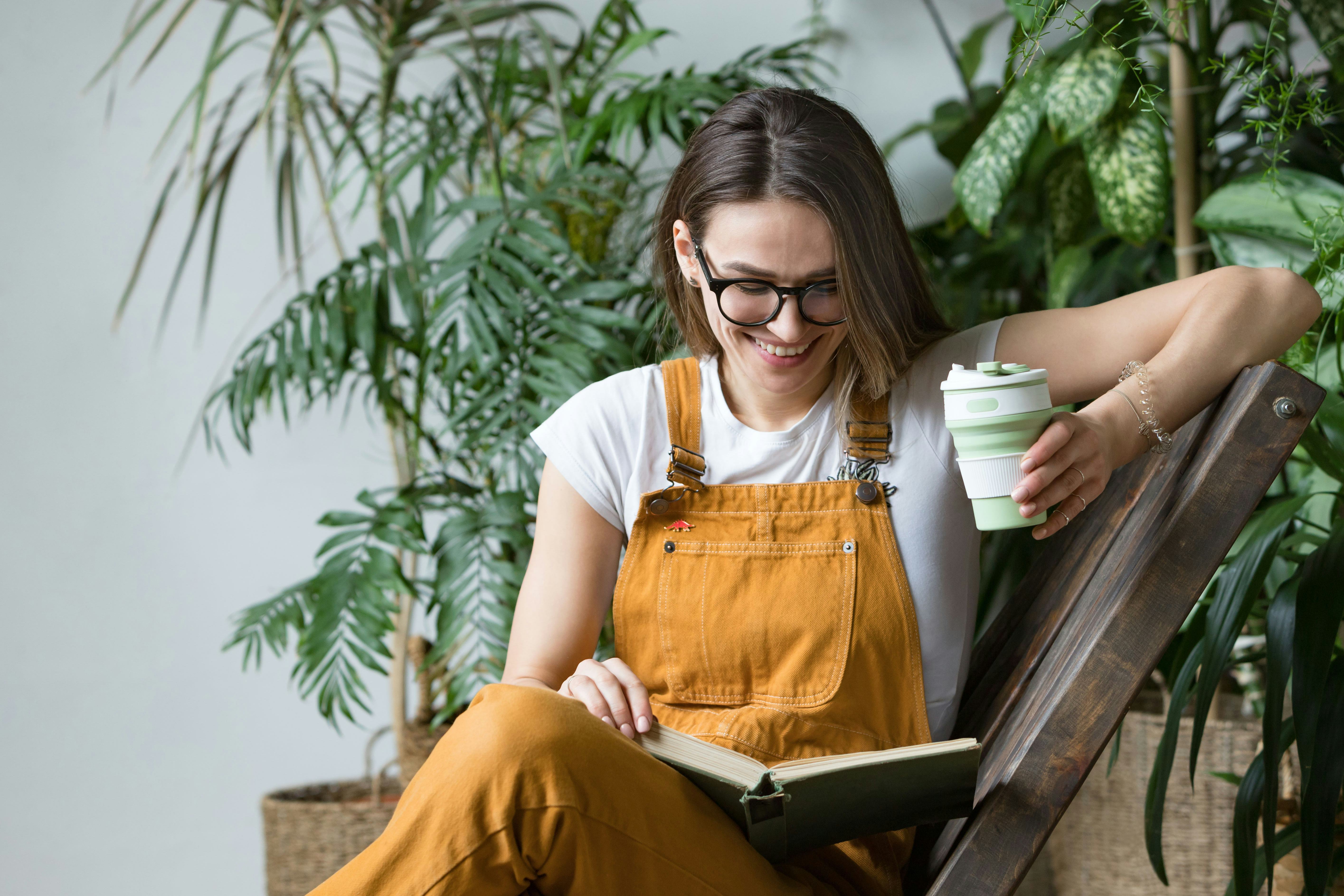 Young female gardener in glasses wearing overalls, resting after work, sitting on wooden chair in ho...