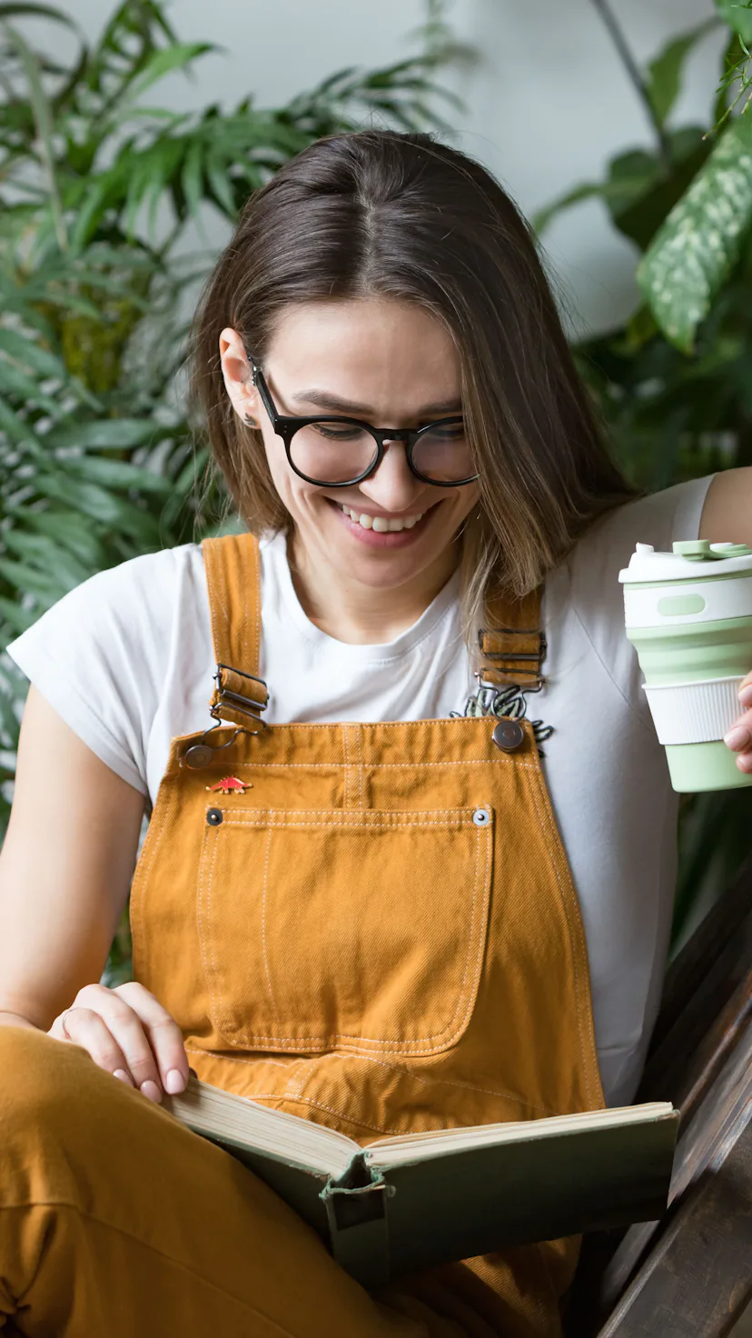 Young female gardener in glasses wearing overalls, resting after work, sitting on wooden chair in ho...