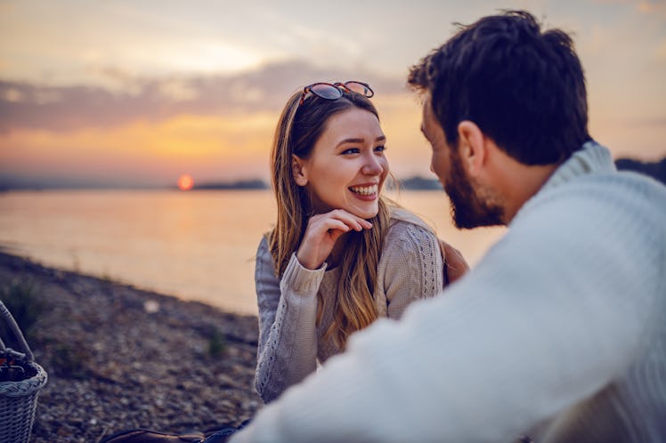 Cute smiling Caucasian brunette sitting on coast near river with her loving boyfriend and flirting. ...
