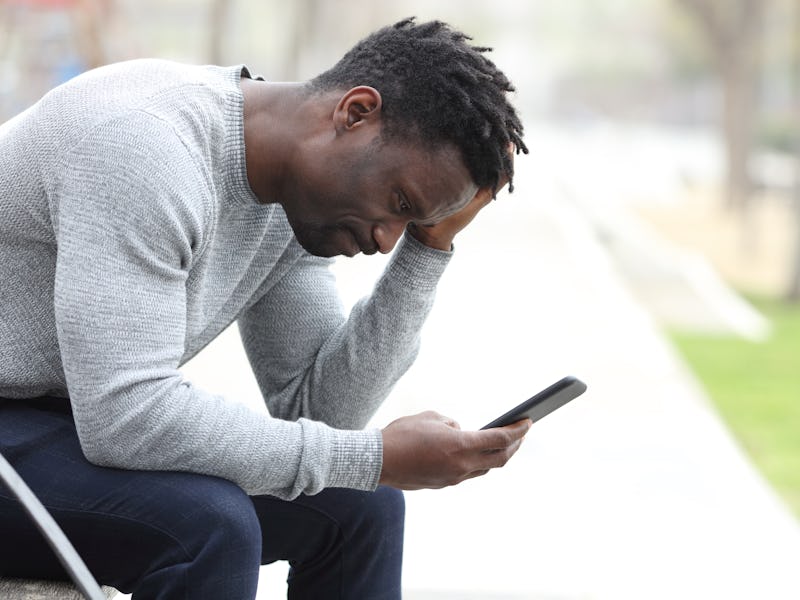 Side view portrait of a sad black man complaining checking mobile phone sitting on a bench in a park