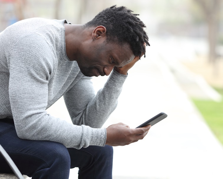 Side view portrait of a sad black man complaining checking mobile phone sitting on a bench in a park