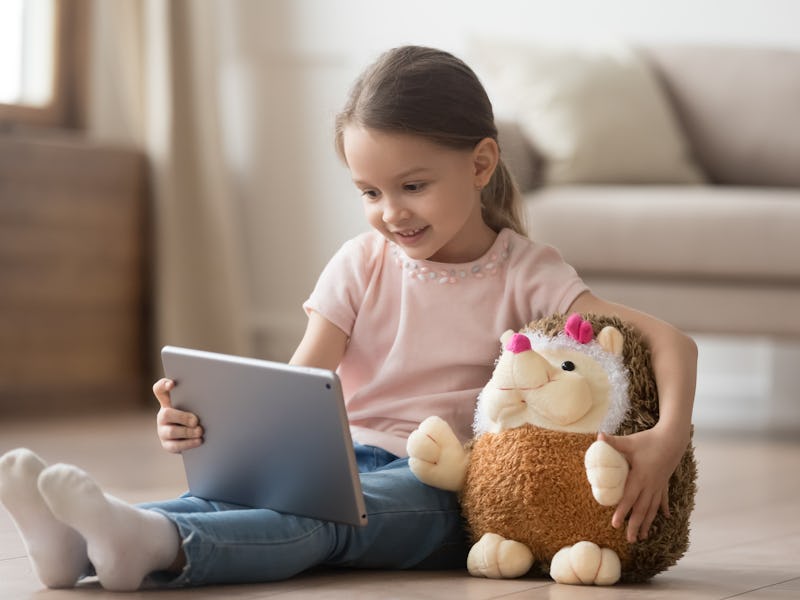 Curious little child girl having fun using digital tablet alone embracing toy sitting on floor, happ...