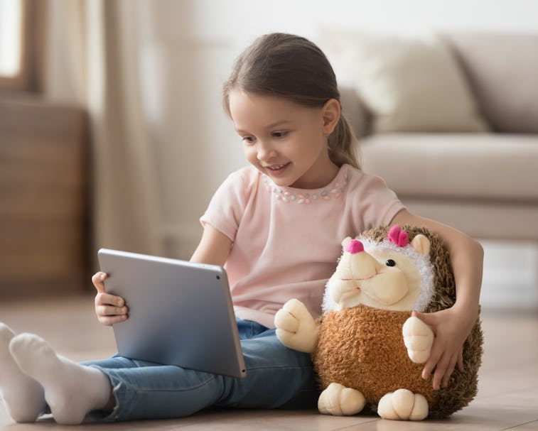 Curious little child girl having fun using digital tablet alone embracing toy sitting on floor, happ...