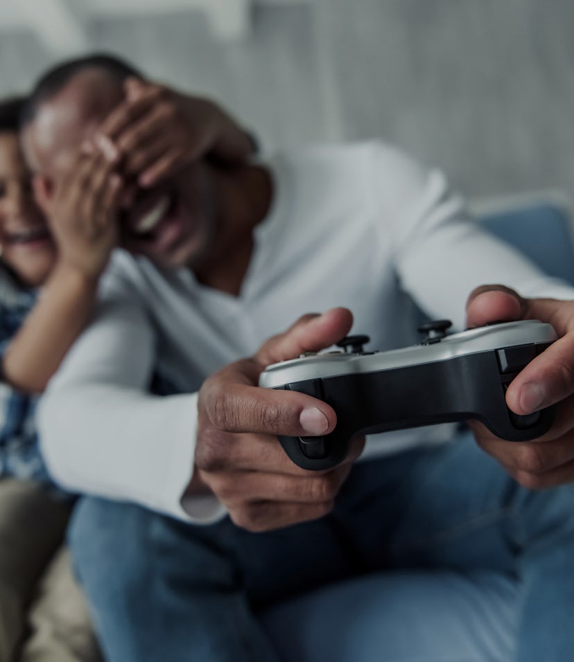 Happy Afro American father and son on couch at home, boy is covering his dad's eyes and smiling whil...