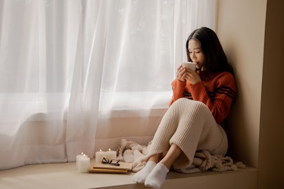 Young beautiful Asian woman holding cup of coffee and marshmallows on top, sitting at home and looki...