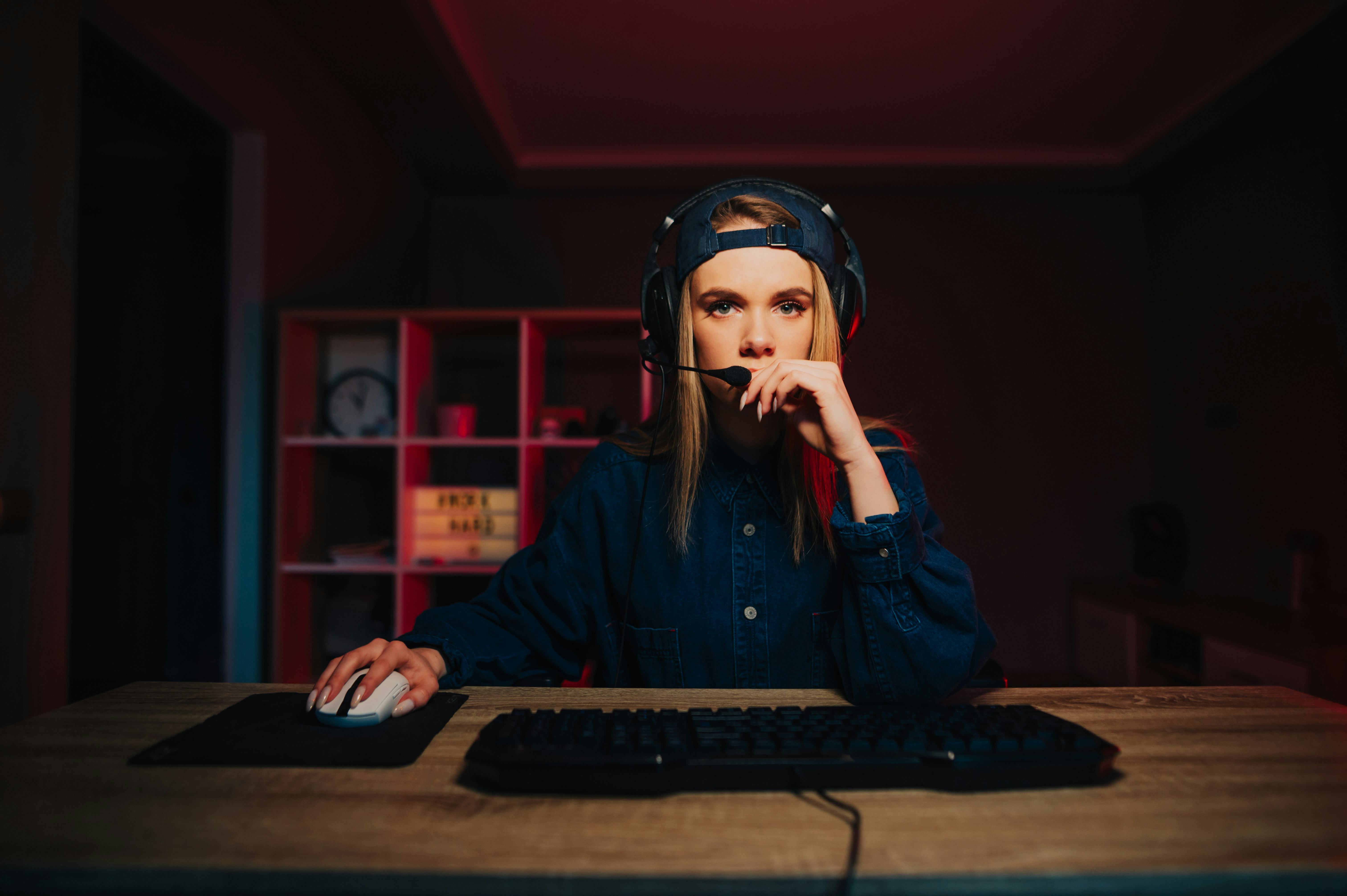 Concentrated woman in casual clothes and headset sitting at the table and playing computer games wit...