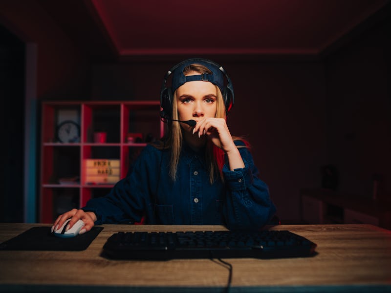Concentrated woman in casual clothes and headset sitting at the table and playing computer games wit...