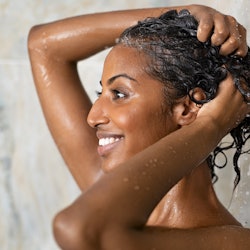 Woman washing hair showering in bathroom at home. Smiling black woman bathing while looking away. Ha...