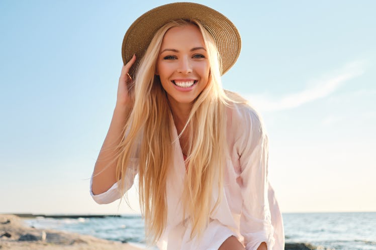 Young attractive smiling blond woman in white shirt and hat joyfully looking in camera with sea on b...