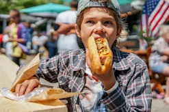 Boy eating barbecue grilled hot dog on family picnic celebrating independence day with flag on the b...