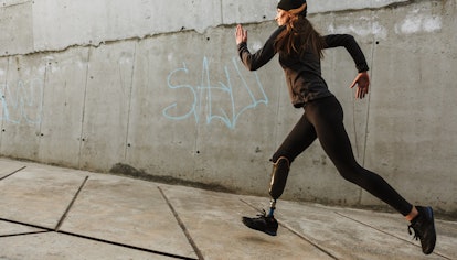 Portrait of disabled athlete woman with prosthetic leg in tracksuit running outdoor along concrete w...