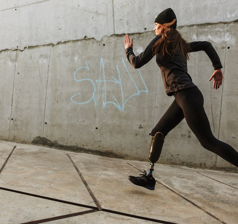 Portrait of disabled athlete woman with prosthetic leg in tracksuit running outdoor along concrete w...