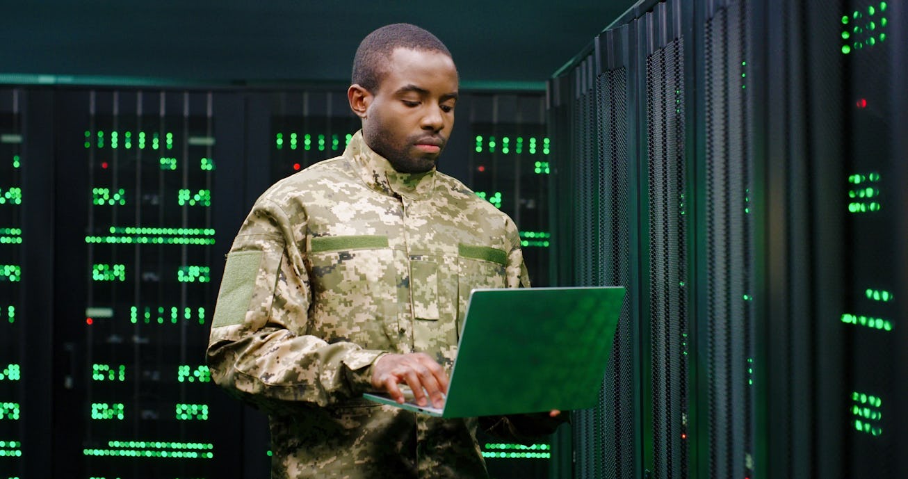 US army male African American officer in uniform stading at server wih secret data and using laptop ...