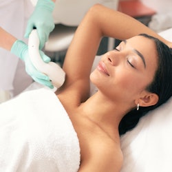 Calm young woman lying with closed eyes and putting on arm up while having laser hair removal proced...