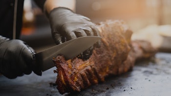 Grill restaurant kitchen. Chef in black cooking gloves using knife to cut smoked pork ribs.