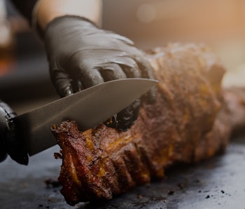 Grill restaurant kitchen. Chef in black cooking gloves using knife to cut smoked pork ribs.