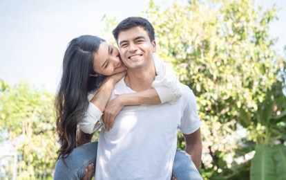 Portrait of cute romantic caucasian guy in checkered shirt, dreamy asian lady rides him on rear. Lei...