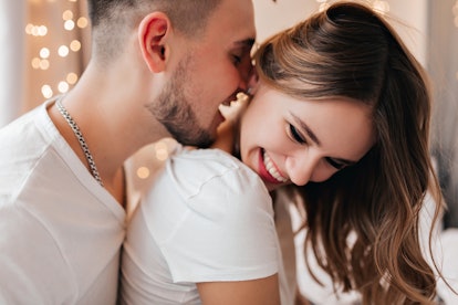 White young woman laughing while husband kissing her neck. Indoor shot of black-haired bearded guy p...
