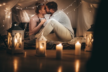 Romantic couple at home sitting on bed together at night with candles lit in the room. Man kissing h...