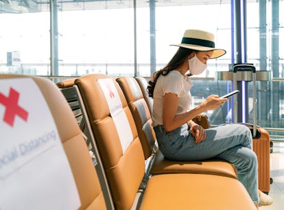 Asian female tourist wearing mask using mobile phone searching airline flight status and sit social ...