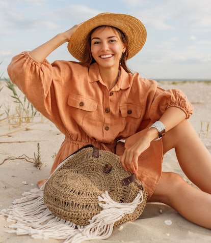 Graceful young  woman in trendy summer linen dress and straw hat posing on the beach near ocean. Bal...