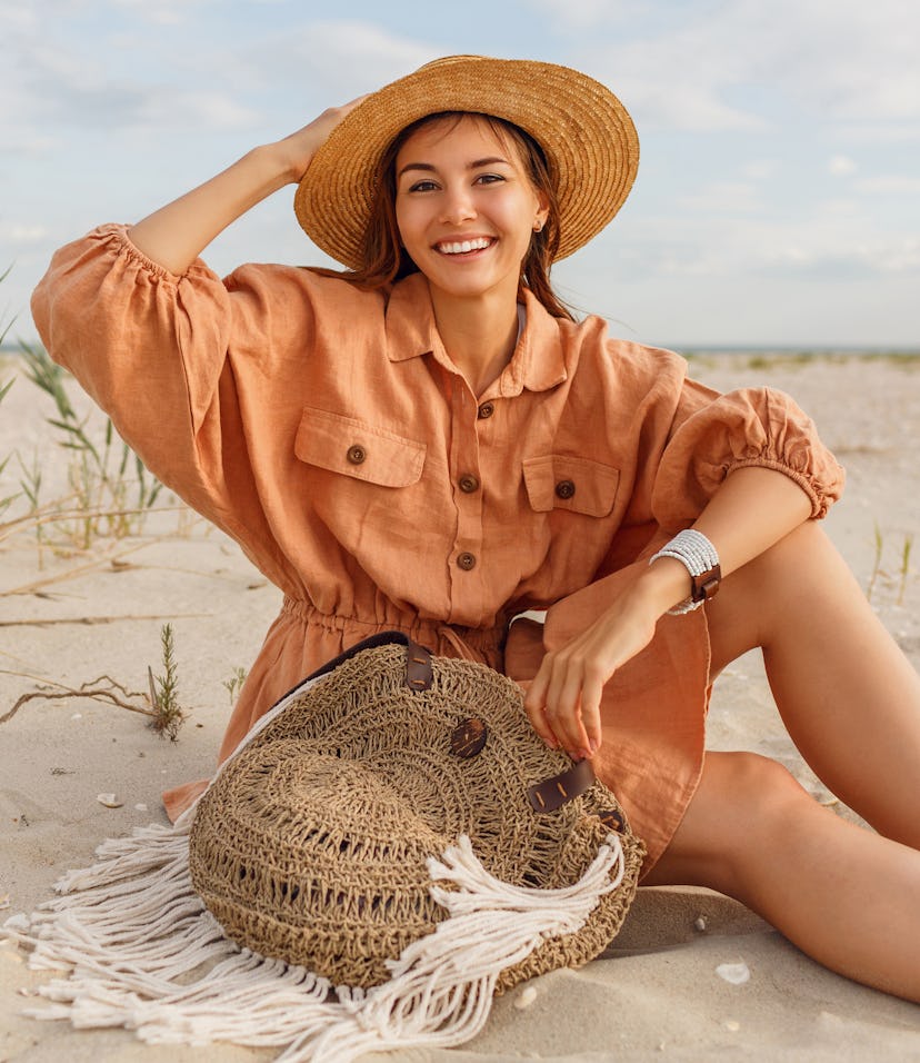 Graceful young woman in trendy summer linen dress and straw hat posing on the beach near ocean. Bal...