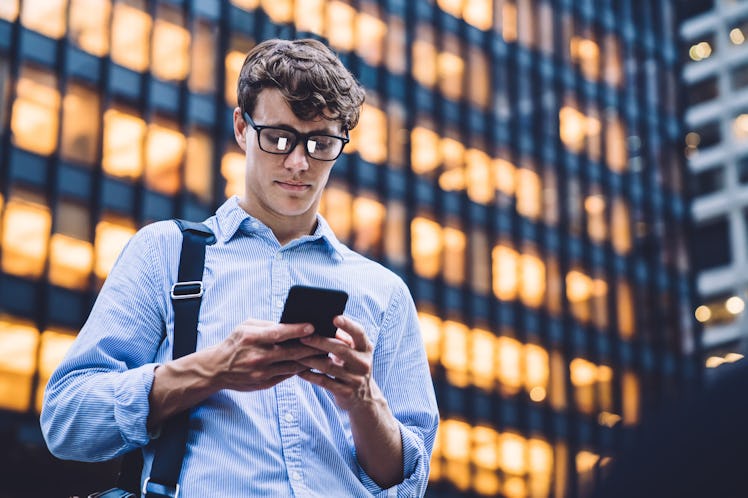 Man in shirt with rolled sleeves and glasses with bag texting on phone standing on background of lig...