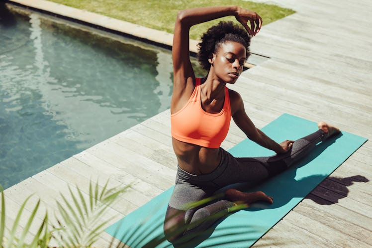 Healthy woman doing yoga stretches by the poolside. African female in sportswear exercising yoga in ...