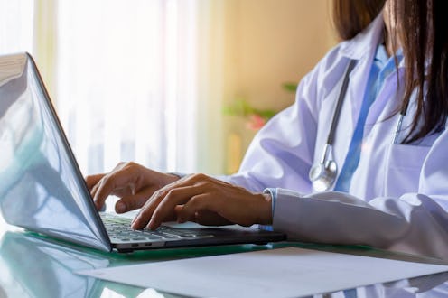 Female doctor in white coat with stethoscope working and typing on laptop computer keyboard at medic...