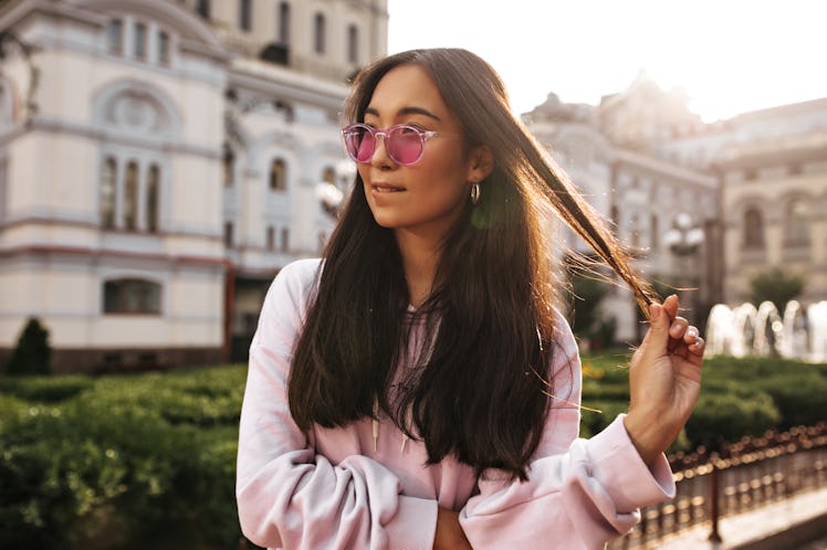 Happy young brunette woman in stylish pink hoodie and sunglasses bites lip, plays hair and poses in ...