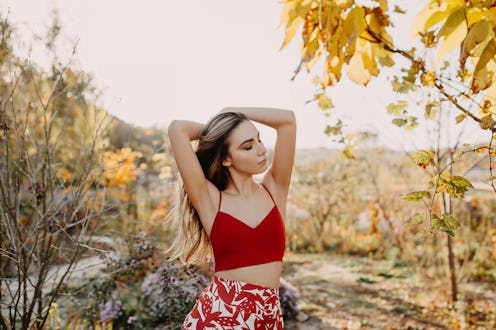 Young woman with long blonde wavy hair, wearing a red crop top and a skirt, in a park.