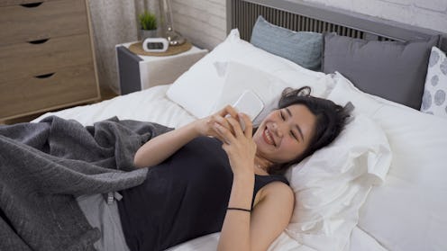 happy lazy asian girl covered with blanket is surfing internet with cell phone while lying in her be...