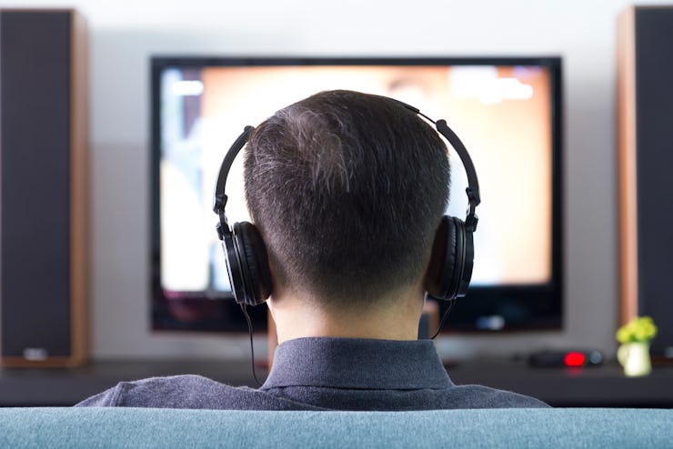 Back side of an Asian man wearing black heaphones in front of blurry out-of-focus television and hom...