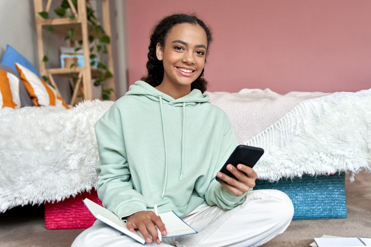 Happy african american teen girl high school student holding cell phone distance learning class usin...